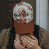 "Mother wearing a vintage floral print trucker hat with embroidered 'MOTHER' lettering, holding her baby."