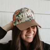 Woman wearing floral YHWH hat, adjusting brim, smiling in outdoor setting.