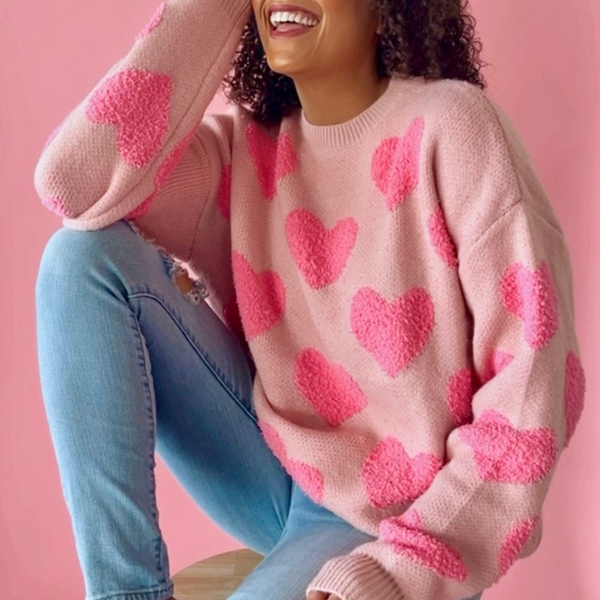 Model sitting in a pink knit sweater with textured heart pattern, paired with ripped jeans, styled against a pink background.