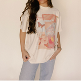 Woman wearing cream-colored “Fullness of Joy” t-shirt with artistic butterfly and text print in pinks and corals, paired with light denim jeans.