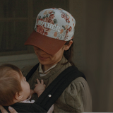"A woman wearing the 'MOTHER' floral hat with a brown bill, smiling while carrying her baby in a sling."