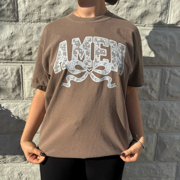 Woman wearing brown Comfort Colors "AMEN" graphic t-shirt with lace-inspired design and bow detail, standing against a stone wall.