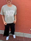 Model wearing sage green Comfort Colors "AMEN" t-shirt, paired with a floral baseball cap, standing against a red brick wall.