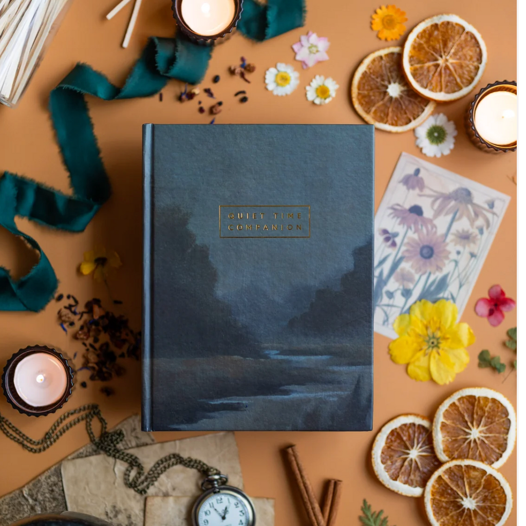 Quiet Time Companion journal with a dark landscape cover and gold foil lettering, surrounded by candles and dried flowers.