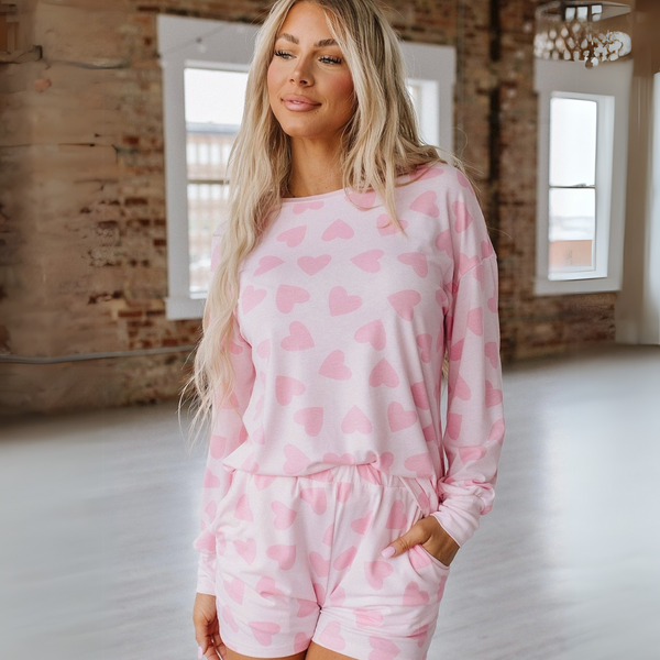 Woman wearing pink Heart Print Loungewear Set, long-sleeve top and shorts, standing in a modern loft.