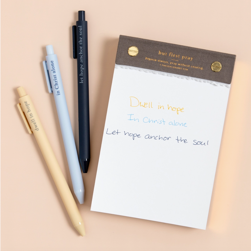 Faith-based pens writing spiritual phrases in ink, laid out on a flat-lay with a notepad.

