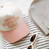 Flat-lay of the "Revival Starts With Family" hat in soft pink and cream, paired with striped shirt and white sunglasses.