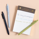 Faith-based pens writing spiritual phrases in ink, laid out on a flat-lay with a notepad.

