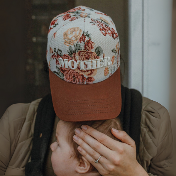 "Mother wearing a vintage floral print trucker hat with embroidered 'MOTHER' lettering, holding her baby."

