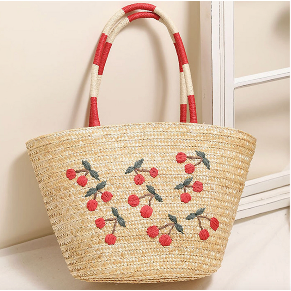 Straw tote bag with embroidered cherries and red striped handles leaning against a white window.

