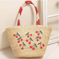 Straw tote bag with embroidered cherries and red striped handles leaning against a white window.

