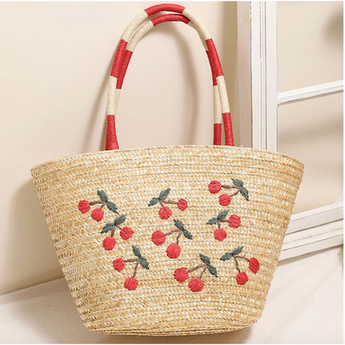 Straw tote bag with embroidered cherries and red striped handles leaning against a white window.

