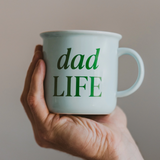 Hand holding a light blue ceramic mug with green “Dad Life” text

