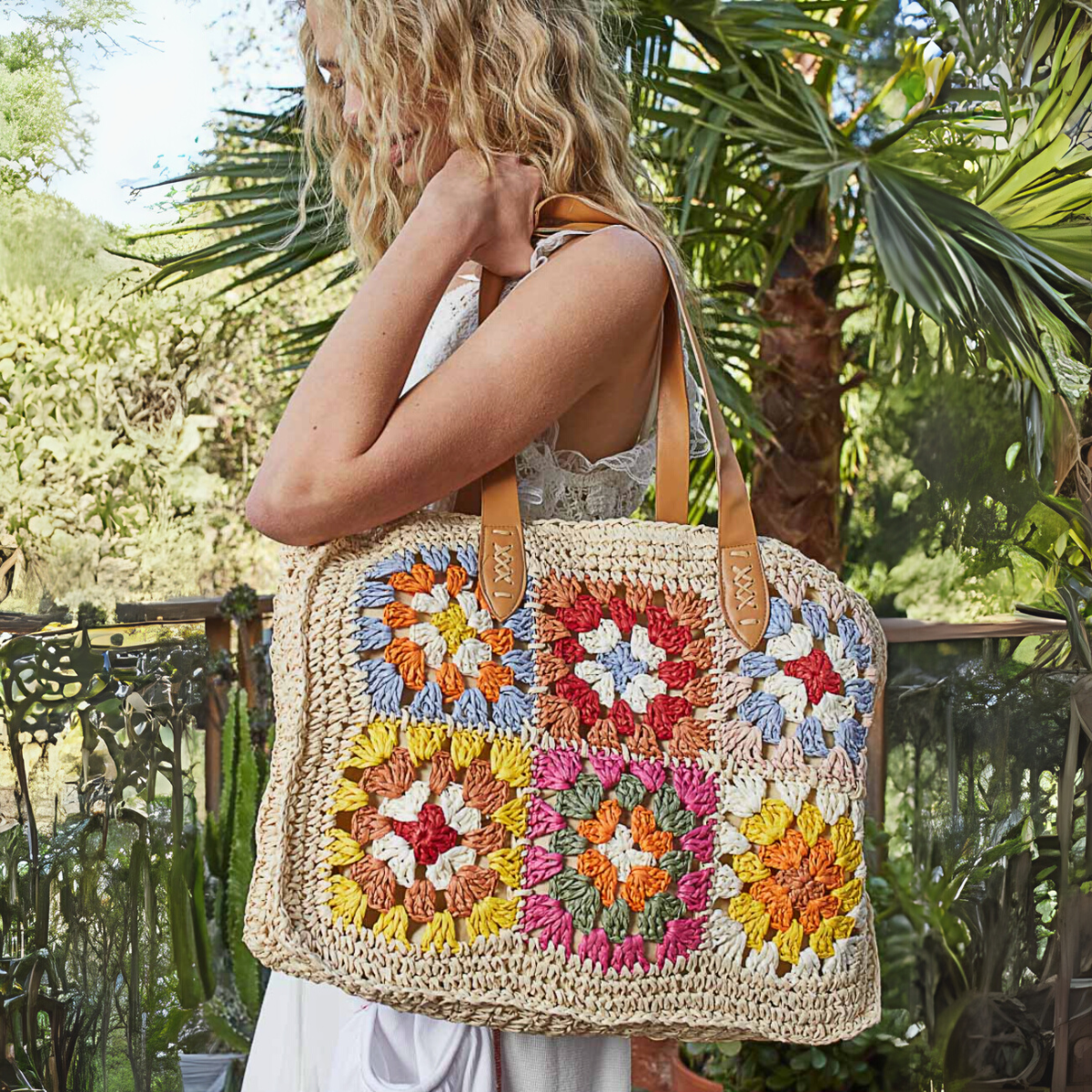 Woman holding a handmade crochet tote bag featuring colorful patchwork squares and leather straps in a natural outdoor setting.