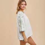 Front view of white oversized jersey tee with green embroidered hop vines on sleeves and pocket detail.