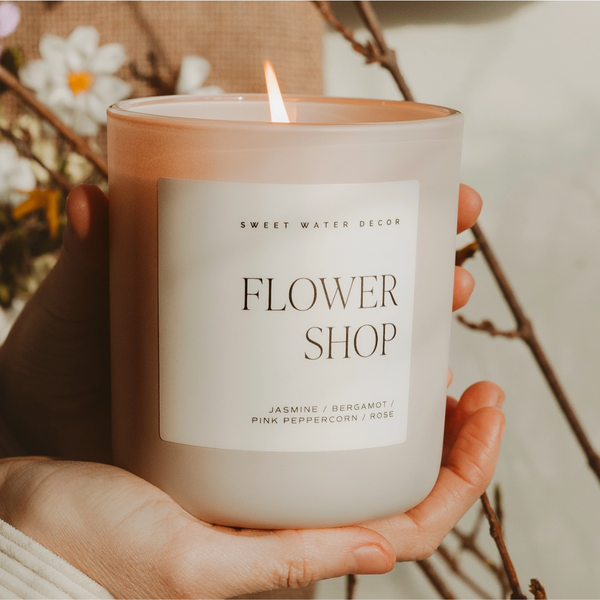 Hands holding a lit Flower Shop candle in a frosted glass jar with white label.

