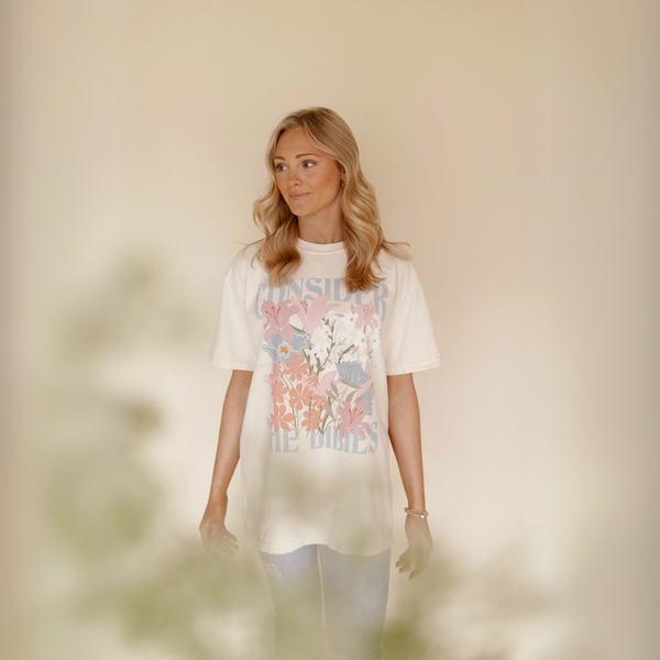 Woman wearing the Consider the Lilies graphic tee, standing with soft floral background.