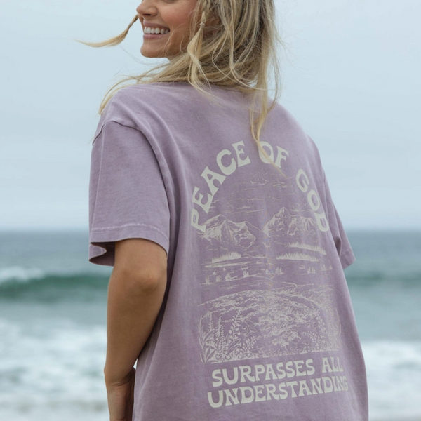 Woman wearing a lavender "Peace of God" graphic t-shirt on the beach, showcasing the back design.