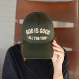 Person holding a forest green 'God Is Good All the Time' baseball cap with bold embroidered lettering."