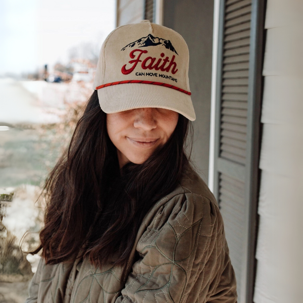 "Woman wearing a beige corduroy 'Faith Can Move Mountains' hat with red rope detail, standing outside a rustic home."

