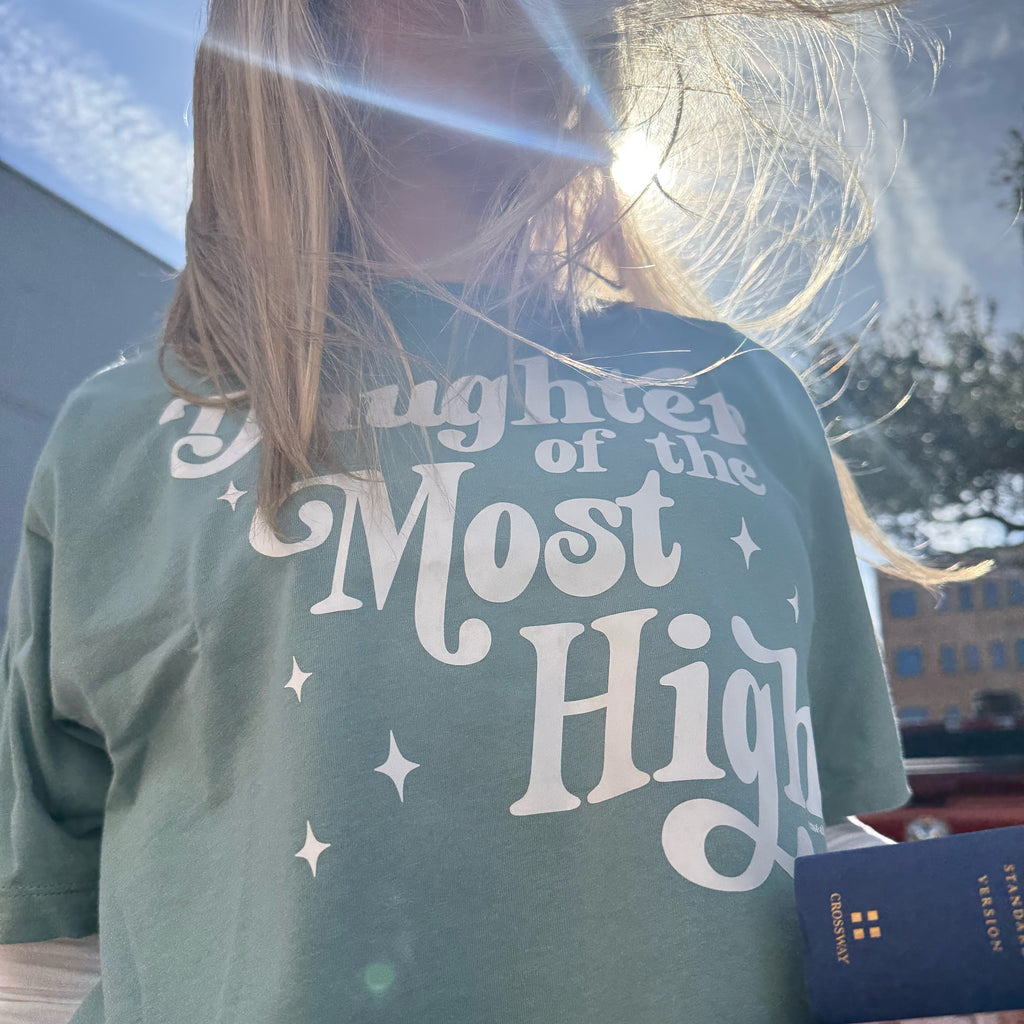 Person wearing a green t-shirt with 'Daughter of the Most High' text, holding books.