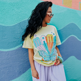 Full view of model in yellow “MAMA” shirt with butterfly design, styled with sunglasses and colorful wall background.