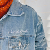 Close-up of the "Elevated Faith" embroidered detail on the front pocket of the denim jacket.