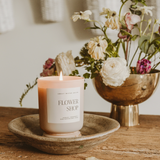 Flower Shop candle burning in a dish with floral arrangement in the background.

