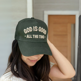 Woman wearing a faith-based trucker hat with 'God Is Good All the Time' embroidery, smiling in a relaxed outdoor setting.