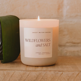 Wildflowers and Salt candle lit on a wooden surface next to a green plant, emitting a warm, relaxing glow.