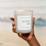 A hand holding the Island Air candle with the ocean in the blurred background, evoking a tropical beach vibe.

