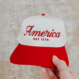 Hand holding red and white embroidered America EST. 1776 baseball cap

