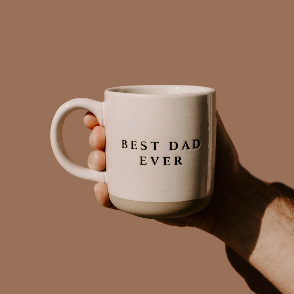 Close-up of hand holding beige stoneware mug with “Best Dad Ever” engraved, against neutral background.
