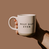 Close-up of hand holding beige stoneware mug with “Best Dad Ever” engraved, against neutral background.