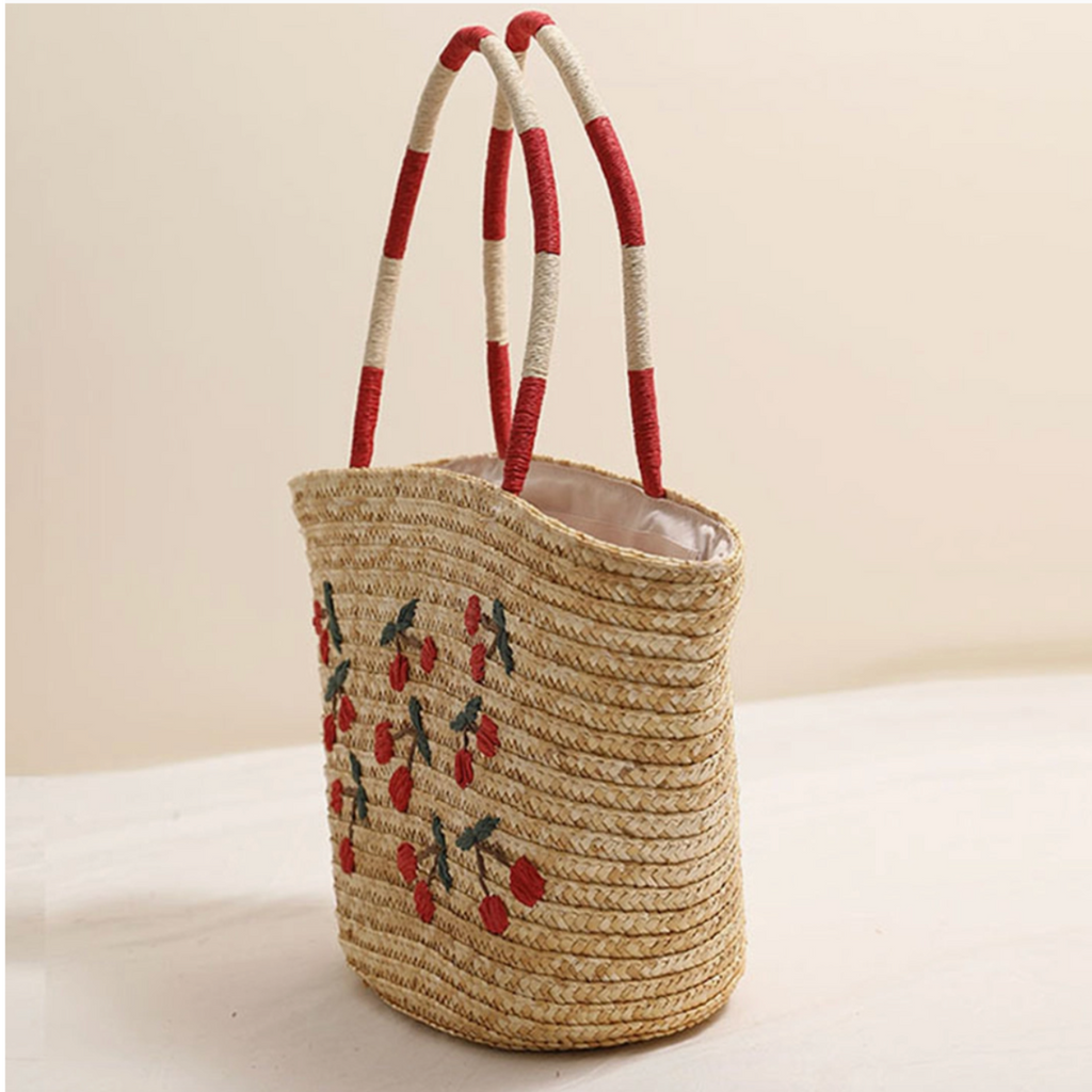 Side angle of cherry-embroidered straw tote bag with tall striped handles.


