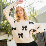 Woman wearing a cream sweater with black bow accents, paired with a black pleated skirt, in a sunny garden setting.