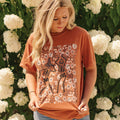 Woman wearing burnt orange Comfort Colors® tee with dancing pumpkin skeletons and floral design, standing in front of white hydrangeas.