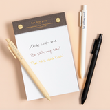 Faith-based pens writing spiritual phrases in ink, laid out on a flat-lay with a notepad.

