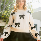 Woman wearing a cream sweater with black bow accents, paired with a black pleated skirt, in a sunny garden setting.