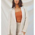 Woman wearing a light-colored blazer, orange top, and beige shorts against a neutral background
