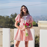 Front view of a textured pink mini dress with bubble sleeves and a white bow tie detail, styled with flowers.