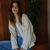 Model wearing the 'It's All Good Washed Jacket' in a soft beige color, styled casually with a gray tank top and blue denim jeans, posed against a wooden background with greenery.