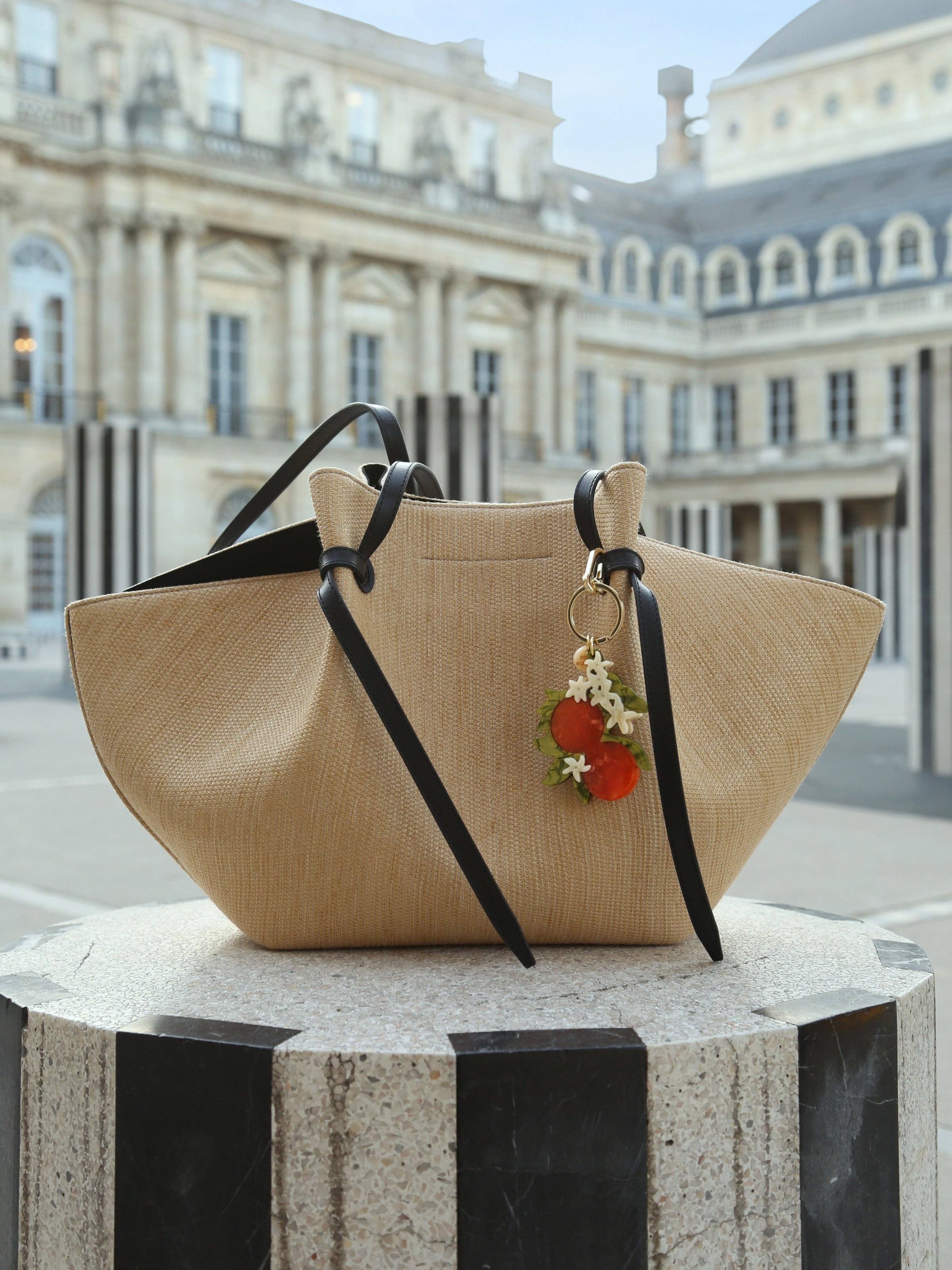Beige handbag with black straps and a floral charm on a pedestal in front of classical architecture.
