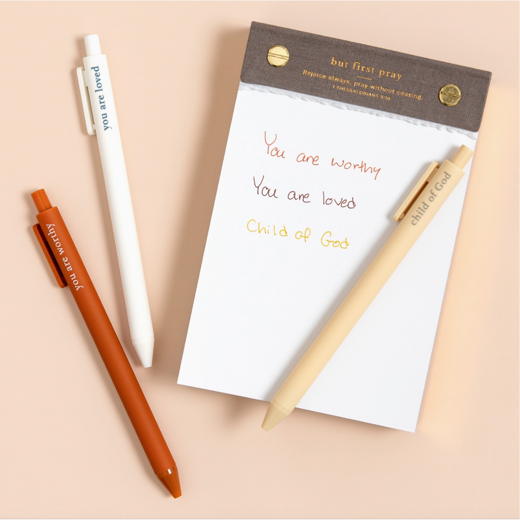 Faith-based pens writing spiritual phrases in ink, laid out on a flat-lay with a notepad.

