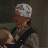 "A woman wearing the 'MOTHER' floral hat with a brown bill, smiling while carrying her baby in a sling."

