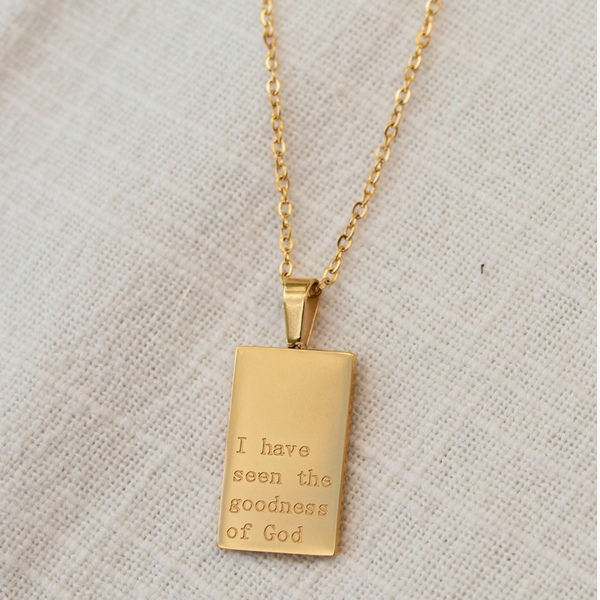 Close-up of gold pendant necklace engraved with “I have seen the goodness of God,” resting on a neutral fabric background.

