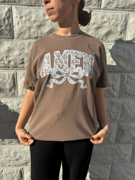 Woman wearing brown Comfort Colors "AMEN" graphic t-shirt with lace-inspired design and bow detail, standing against a stone wall.