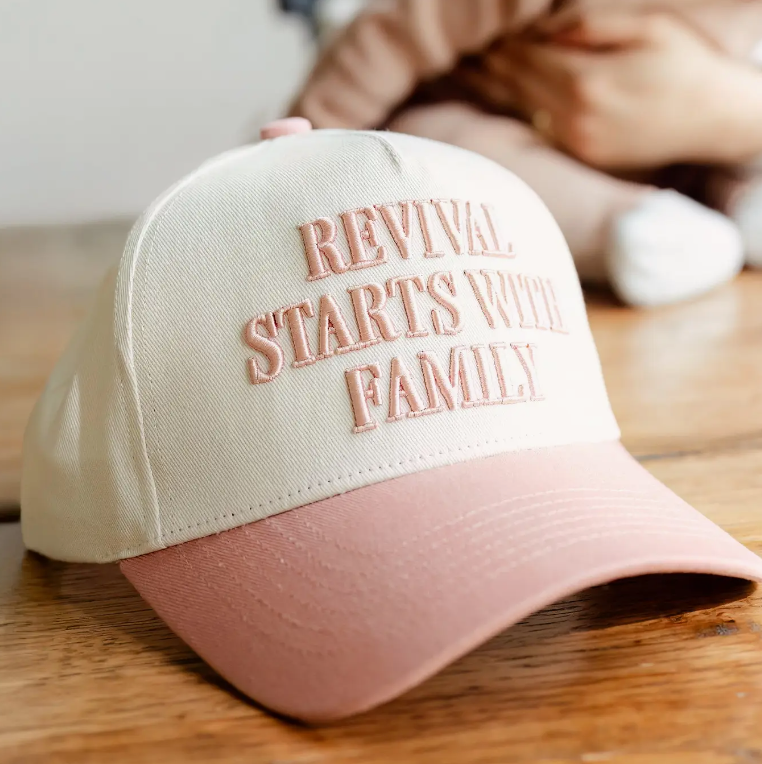 Beige and pink Christian cap with "Revival Starts with Family" text, available at a Lakeland, Florida clothing store, sitting on a wooden table