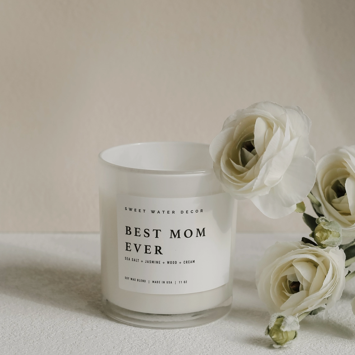 Minimalist white “Best Mom Ever” soy candle with black text, surrounded by white flowers – 11oz jar with sea salt, jasmine, wood, and cream scent.

