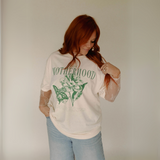 Woman wearing oversized cream-colored “Motherhood” graphic tee with green floral and butterfly design, paired with light-wash jeans.

